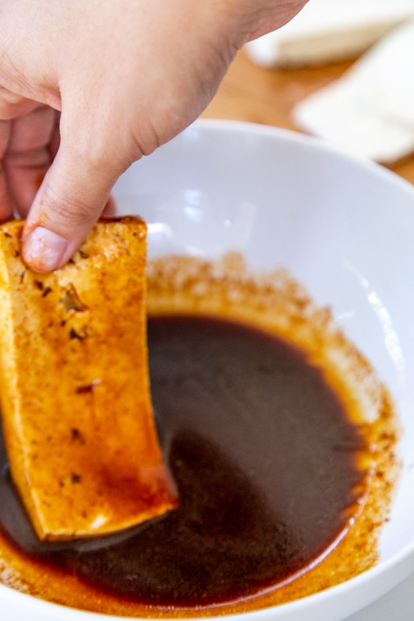 A hand holding a strip of tofu with marinade on it over the bowl of marinade.