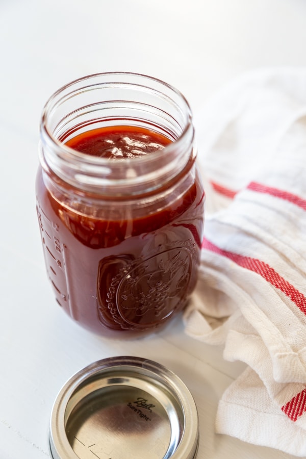A mason jar filled with bbq sauce and a white and red towel next to the jar.