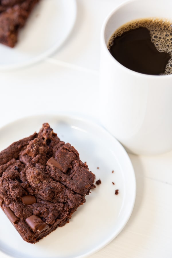 A brownie with chocolate chunks on a white plate with a mug of coffee and another plate with a brownie on a white wood table.