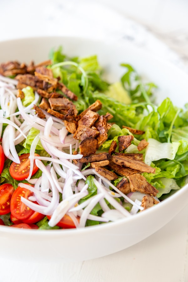 A bowl of chopped lettuce, sliced tomatoes, slivered red onion, and pieces of vegan bacon.