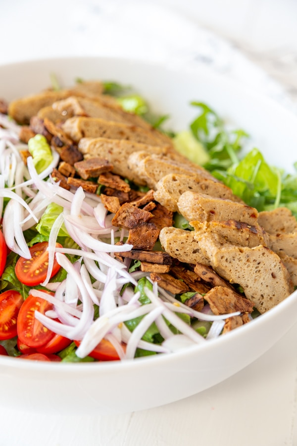 A bowl with chopped lettuce, sliced tomatoes, slivered onion, crumbled bacon, and sliced seitan chicken.