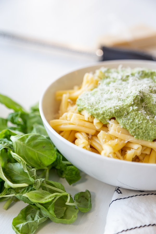 A white bowl with pasta and topped with pesto and basil leaves next to the bowl.