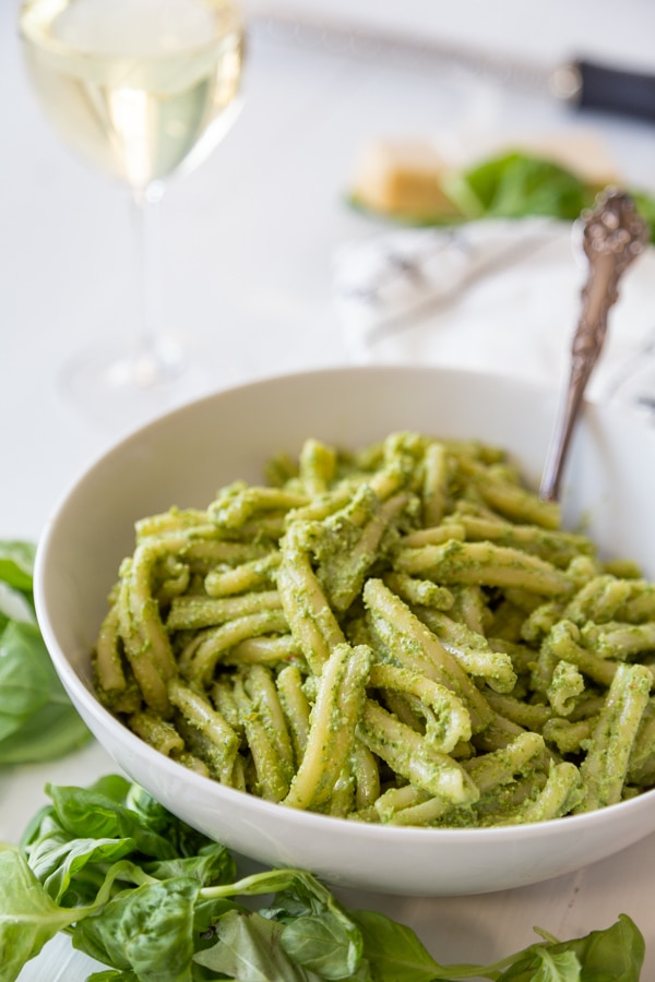 A white bowl with pasta and pesto with a silver fork in the bowl and basil leaves around it.