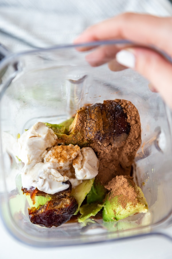A blender with avocado, dates, coconut milk, cocoa powder, and cinnamon.