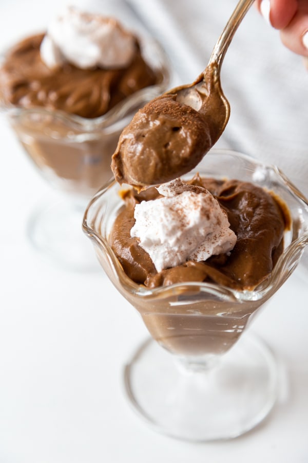 A spoonful of chocolate pudding held over the dish of pudding with whipped topping and another dish of pudding in the background.