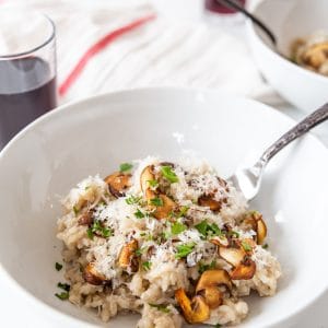 A white bowl with mushroom risotto with a fork in the bowl and a glass of wine and a white and red towel in the background.