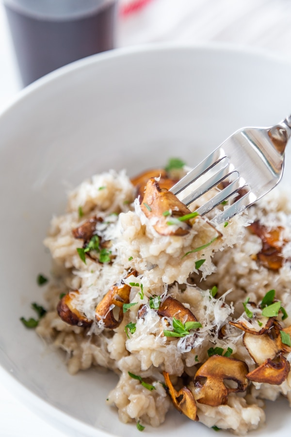 A fork full of mushroom risotto and parmesan cheese over a white bowl of the risotto.