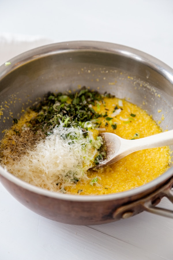 A copper saucepan with polenta and chopped herbs and grated parmesan with a wooden spoon.