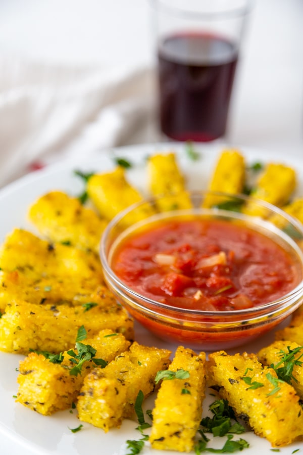 A white plate with a bowl of marinara sauce and a circle of polenta fries around it with a glass of red wine in the background.