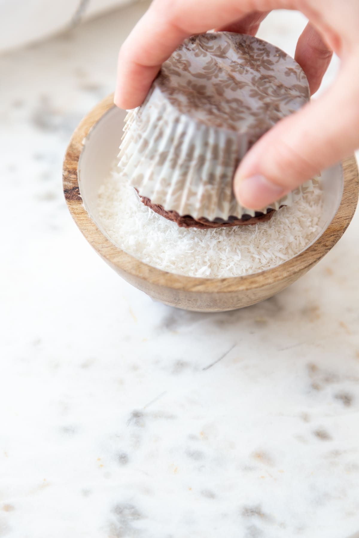 A hand dipping a chocolate frosted cupcake into a bowl of shredded coconut.
