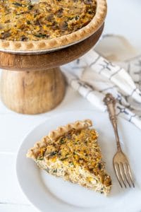 A slice of vegetable quiche on a white plate with a silver fork and a wooden cake stand with the entire quiche behind it.