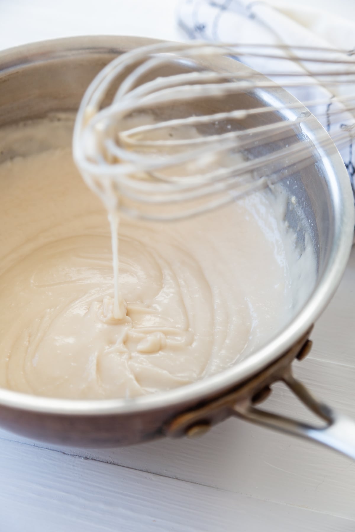 A whisk held over a copper pot of vanilla pudding with pudding dripping off of the whisk.