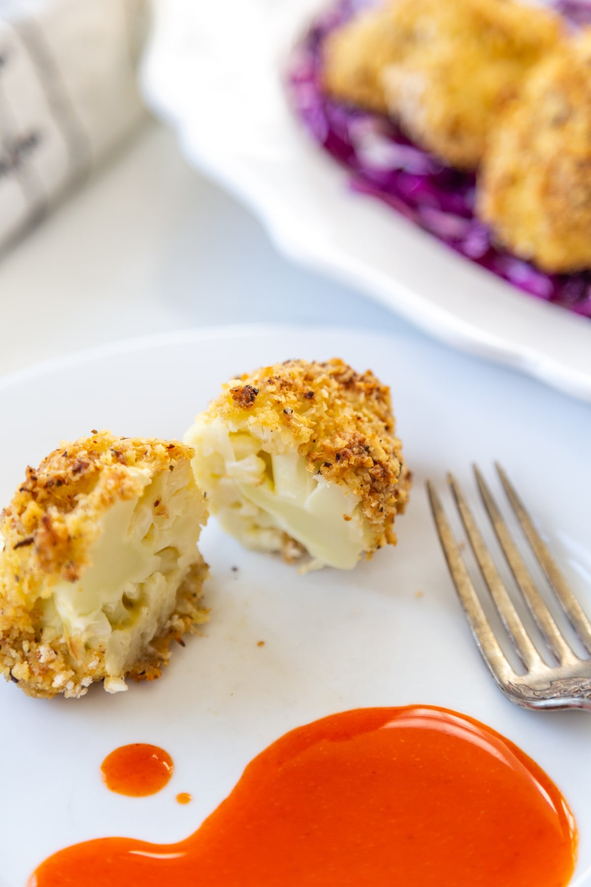 Crispy breaded cauliflower floret cut in half on a white plate with a silver fork and hot sauce on the plate, and a white platter of the cauliflower on a bed of red cabbage in the background.