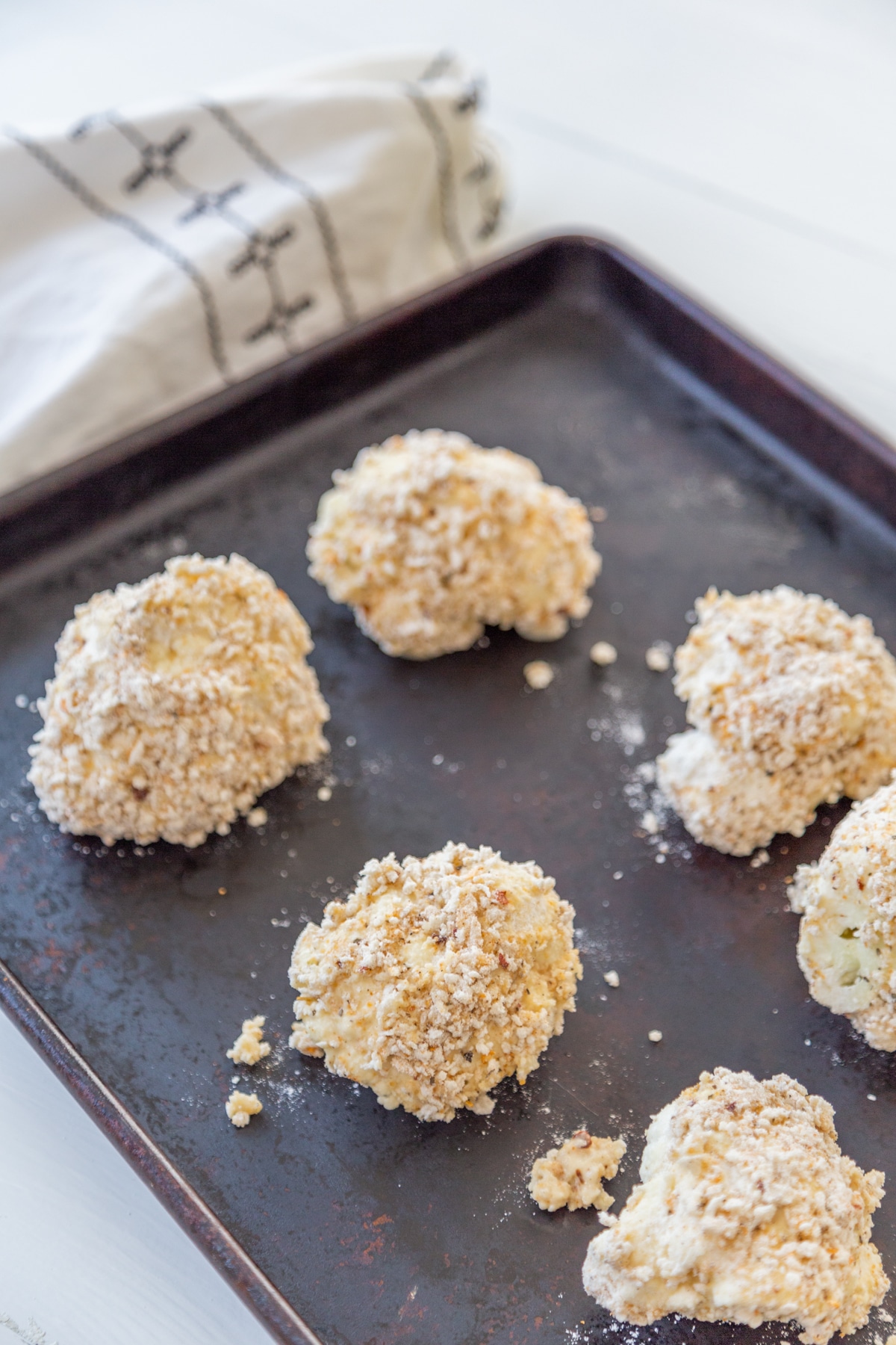 Unbaked cauliflower florets coated in flour and breadcrumbs on a cookie sheet with a white and black towel in the background.