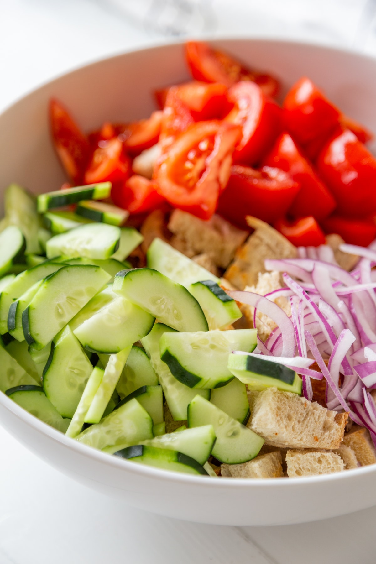 A large white bowl filled with croutons, sliced cucumber, tomatoes, and red onion.