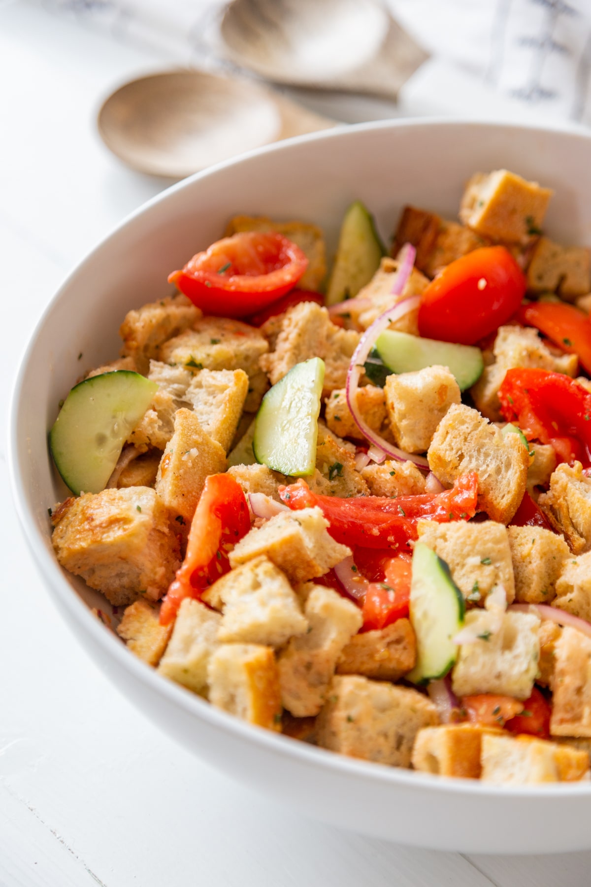A large white bowl with Panzanella salad and white and wood serving spoons in the background.
