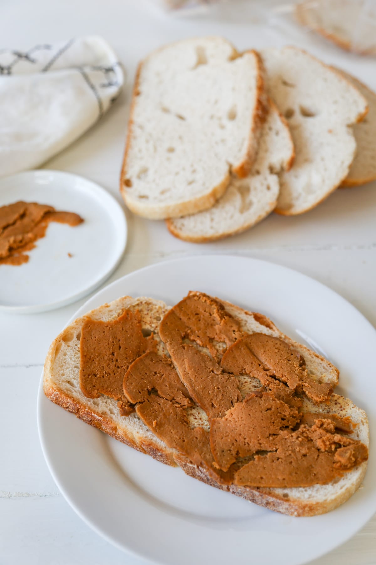 A slice of bread with mustard and sliced ham on a white plate with bread slices and sliced ham in the background.