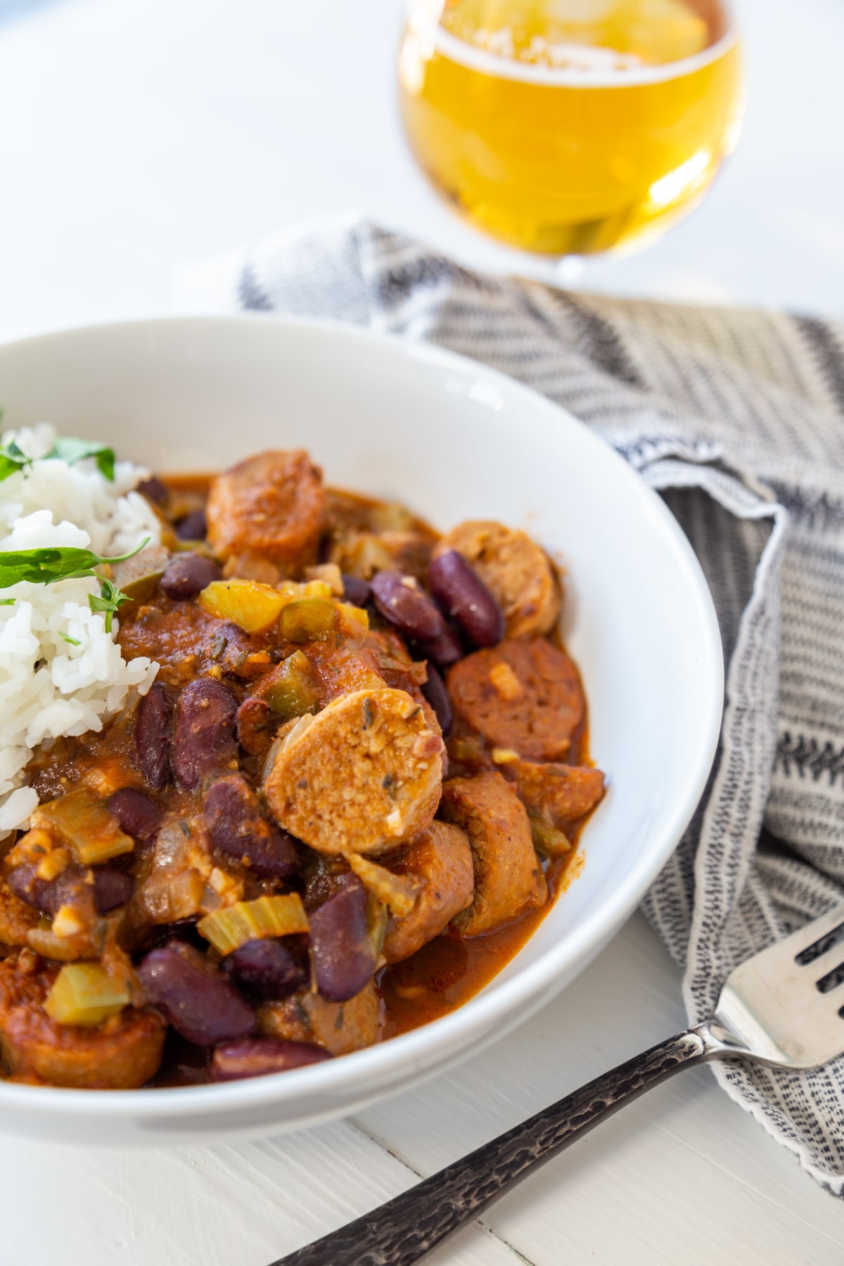 A white bowl filled with sausage and red beans and rice with a silver fork and a black and white towel next to it and a glass of beer in the background.