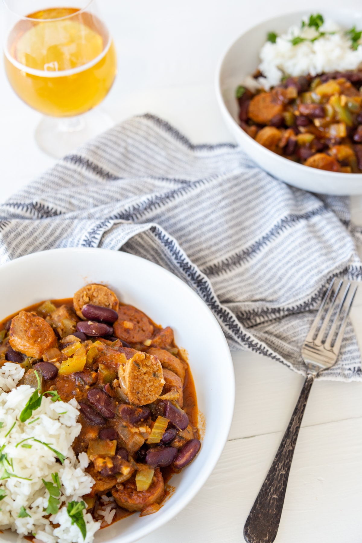 A white table with 2 white bowls of red beans and rice with sausage, and a glass of beer. 