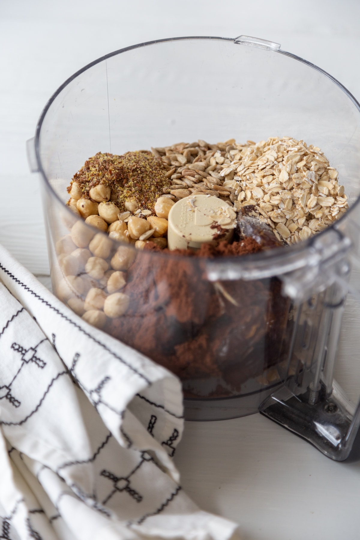 A food processor filled with rolled oats, dates, cocoa powder, and hazelnuts.