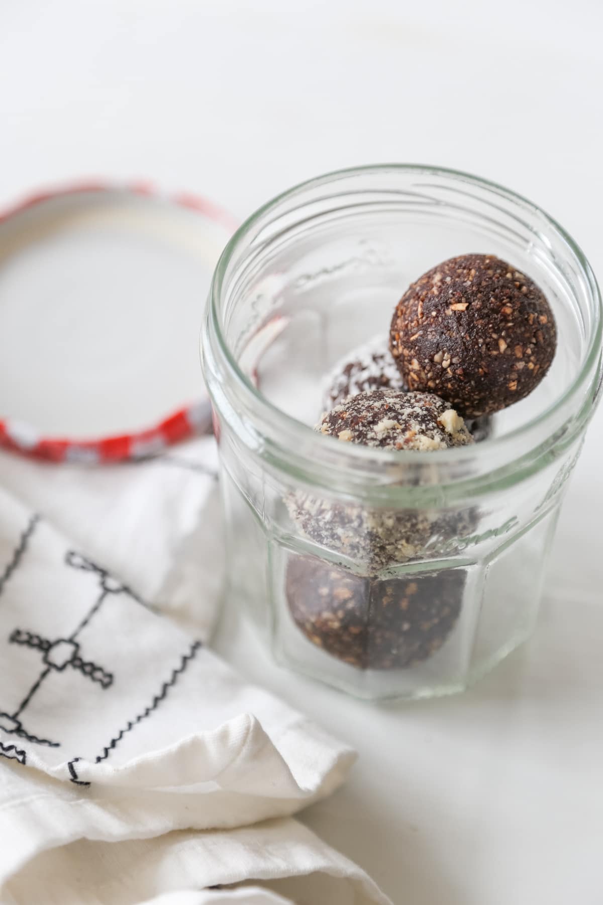 Three chocolate energy balls in a glass jelly jar with the red and white checked lid and a white and black towel next to the jar.
