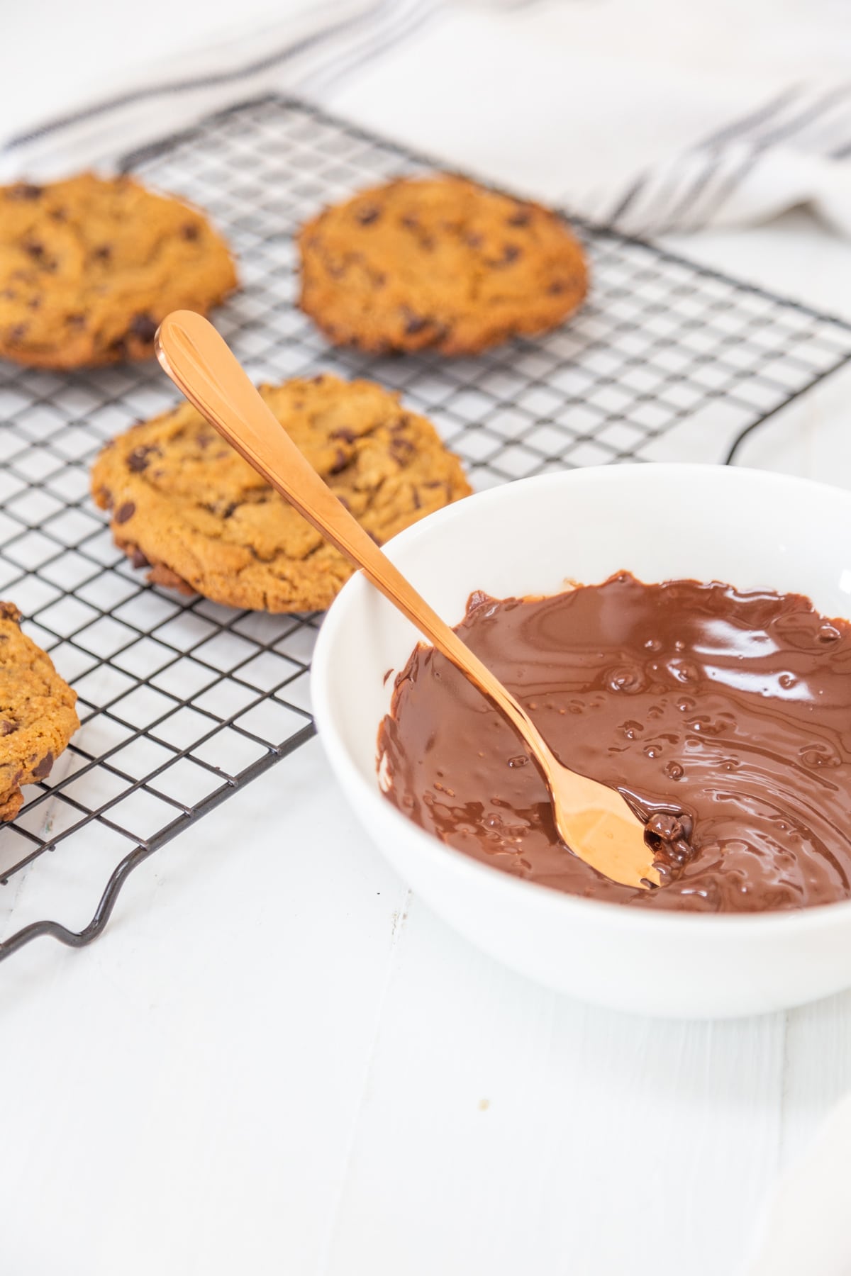 A white bowl of melted chocolate with a copper spoon next to a cooling rack with chocolate chip cookies.