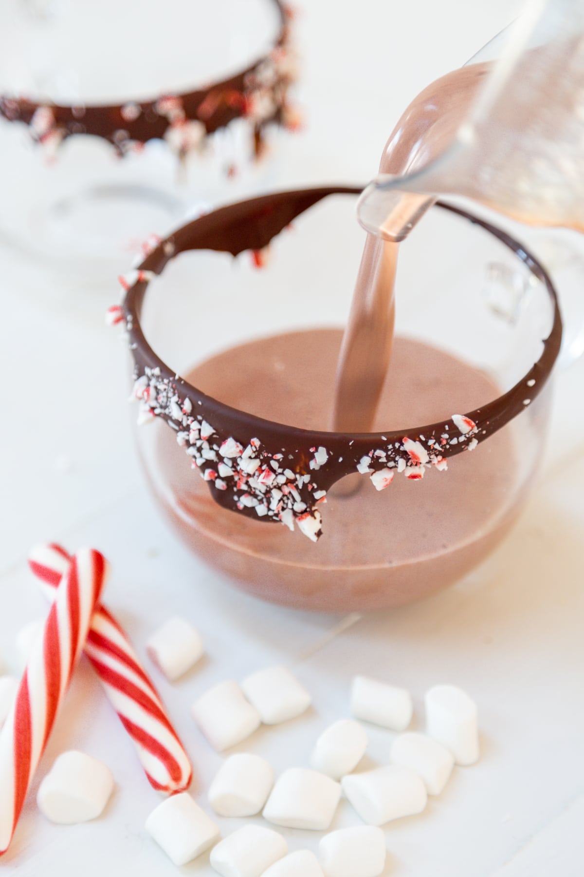 Hot chocolate being poured into a clear glass mug with a chocolate and peppermint rim.