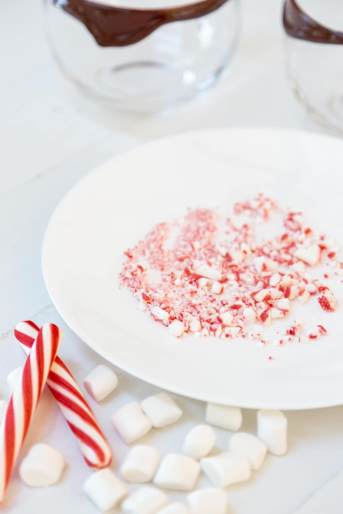 A white plate with crushed candy cane and peppermint sticks and marshmallows scattered next to it, and 2 glass mugs rimmed with chocolate.