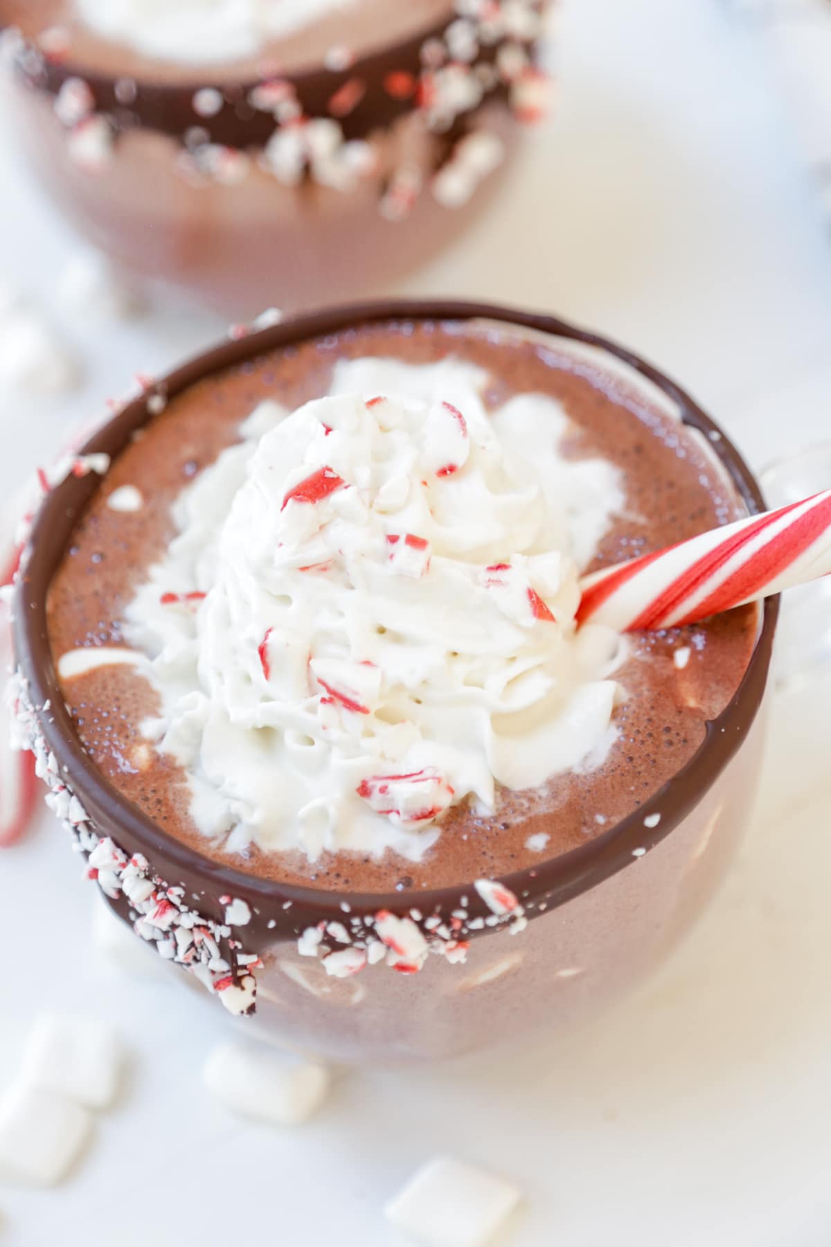 An overhead picture of a clear glass mug with a chocolate & peppermint lined rim filled with hot chocolate and whipped cream, with a peppermint stick in the mug.