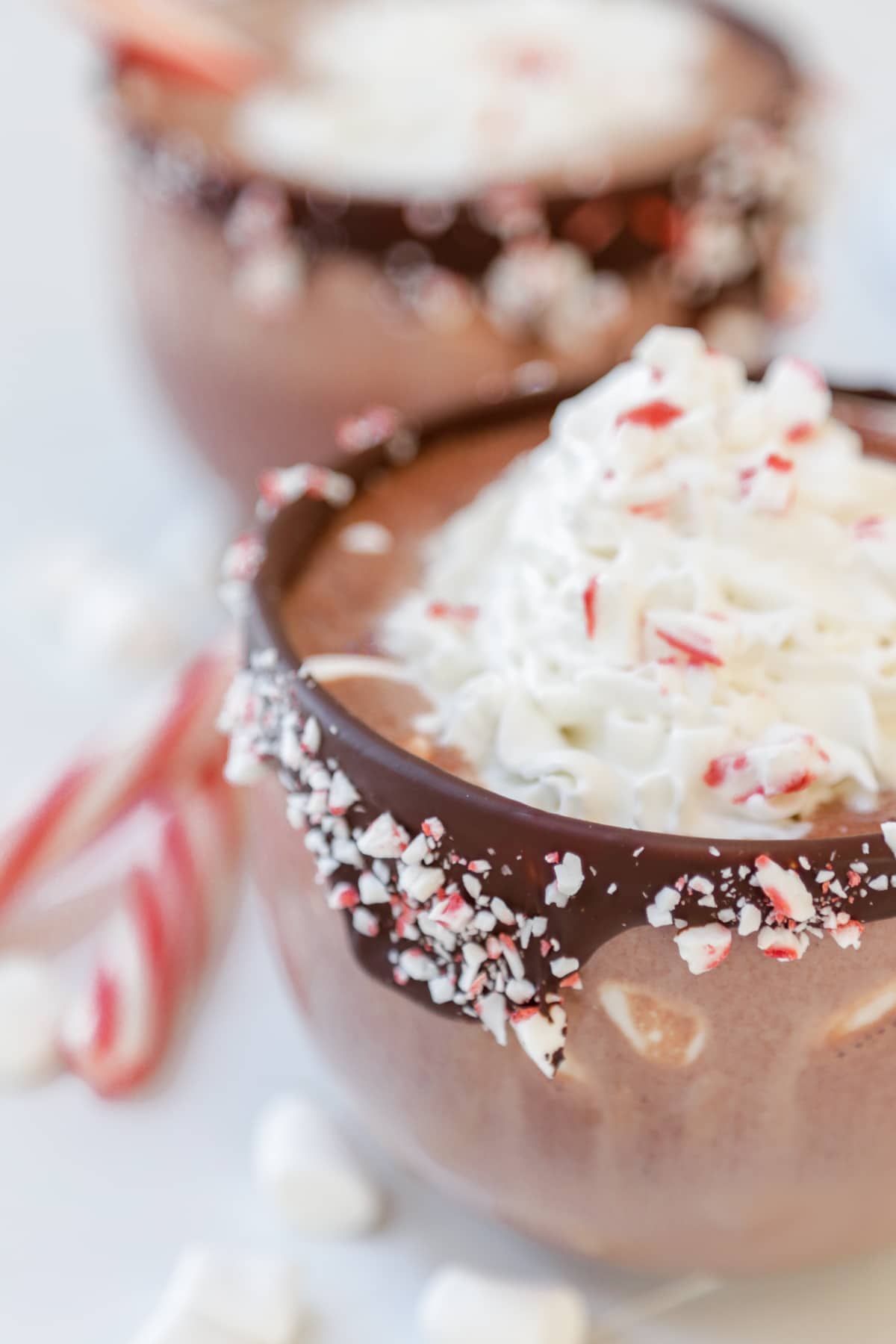 Two clear glass mugs of hot chocolate with whipped cream and a sprinkle of crushed peppermint. The rims of the mugs are dipped in chocolate and crushed peppermint.