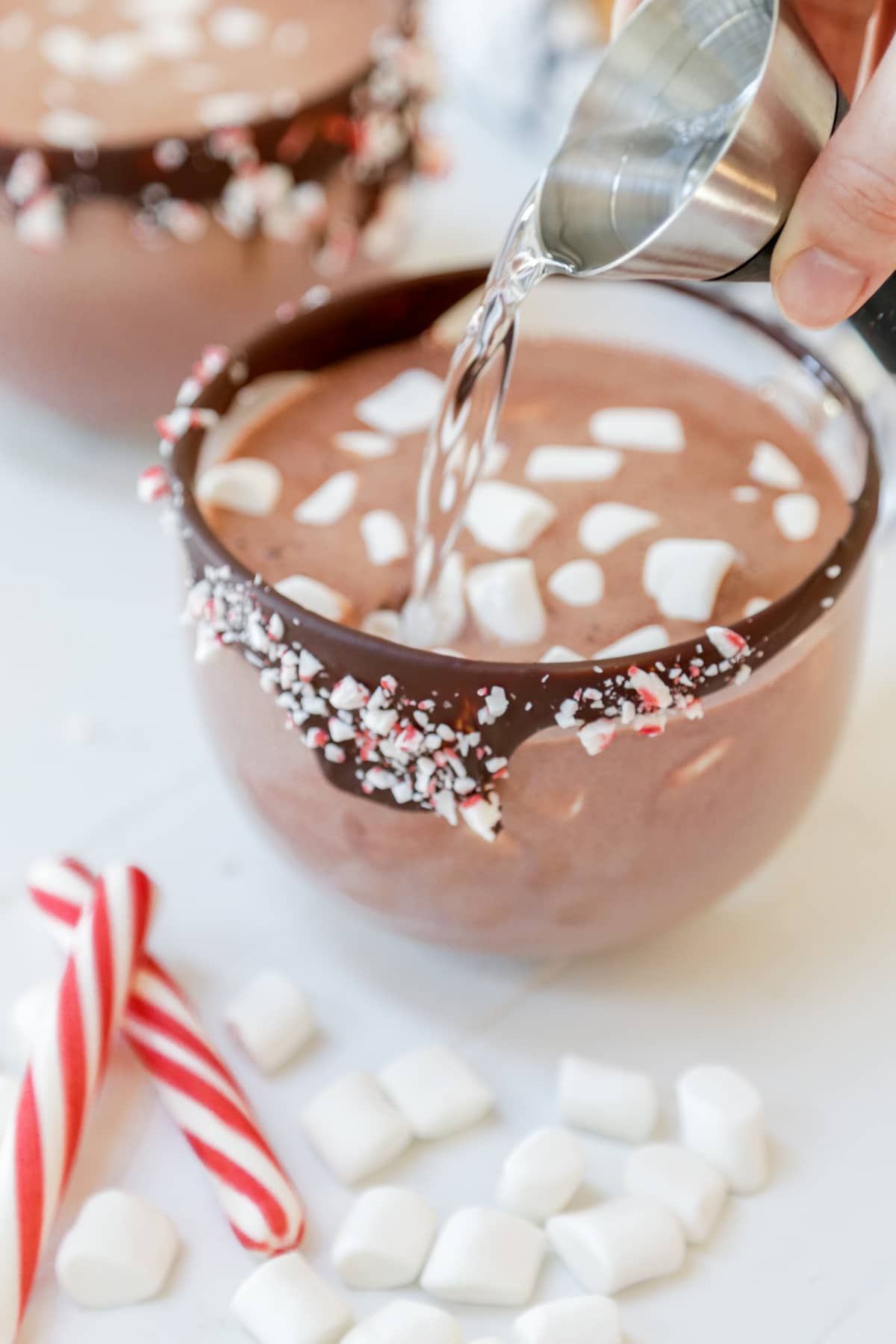 A hand pouring a shot of clear liquid into a clear glass mug rimmed in chocolate and crushed peppermint filled with hot chocolate and marshmallows.