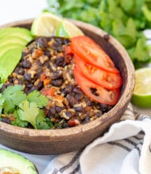 A brown wood bowl with black beans and rice with vegetables and an avocado and cilantro around the bowl.