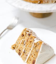 A slice of carrot cake with cream cheese frosting on a white plate with the whole cake in the background.